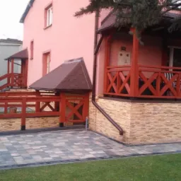 Ułożona kostka brukowa na podjeździe przed domem z drewnianą pergolą i balustradą w kolorze czerwonym.