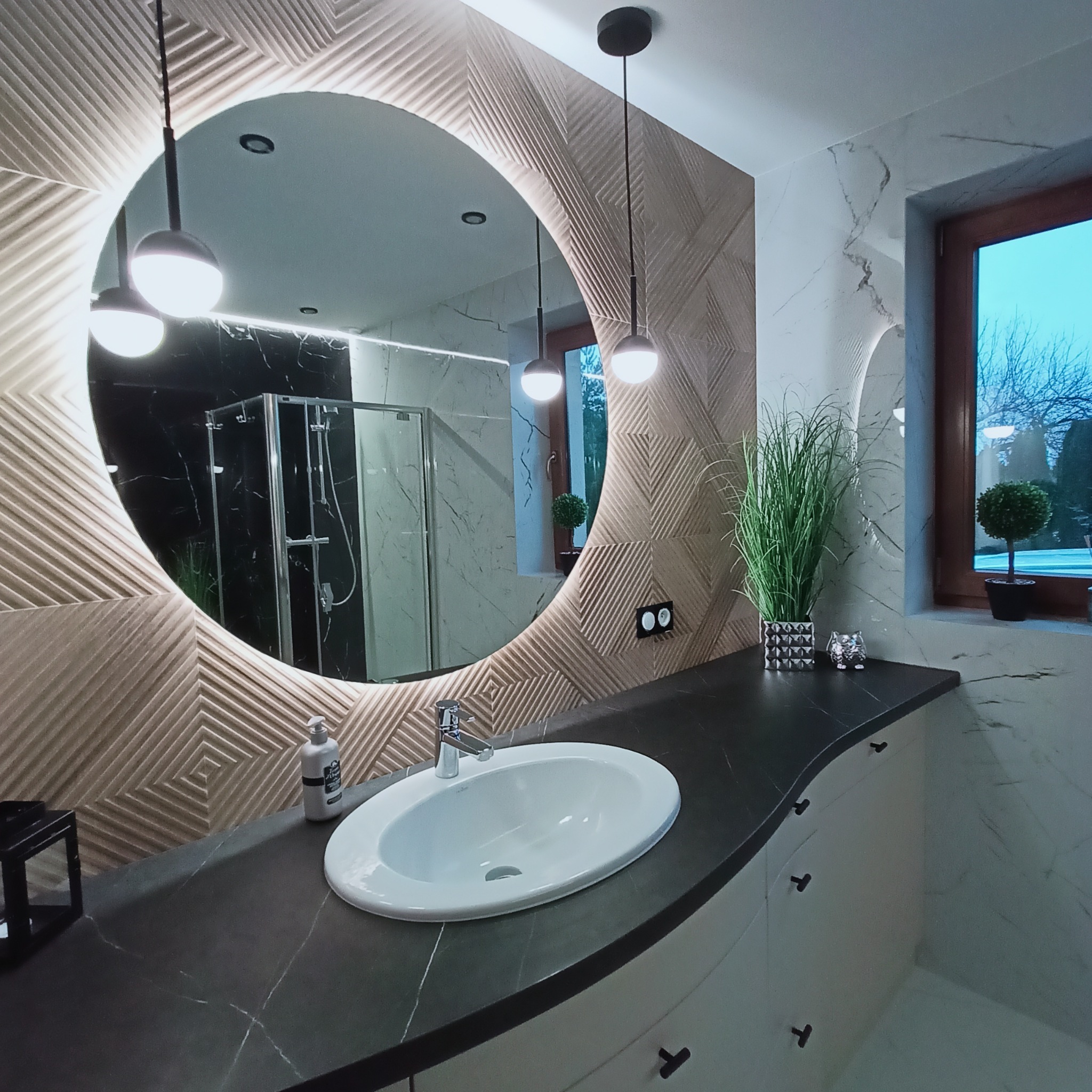 Nowoczesna łazienka z okrągłym lustrem podświetlanym LED, geometryczną mozaiką na ścianie, czarnym blatem i umywalką wpuszczaną, oświetlona wiszącymi lampami kulami i z widokiem na ogród przez okno.
