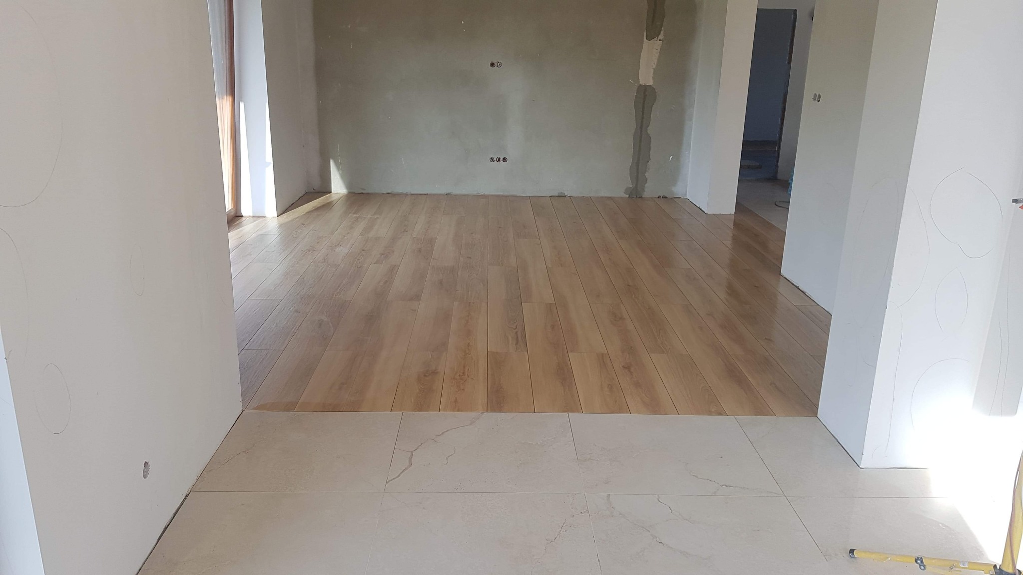 Połączenie jasnych, marmurkowych płytek podłogowych i drewnopodobnej podłogi w surowym wnętrzu, widoczne ślady po pracach wykończeniowych na ścianach.