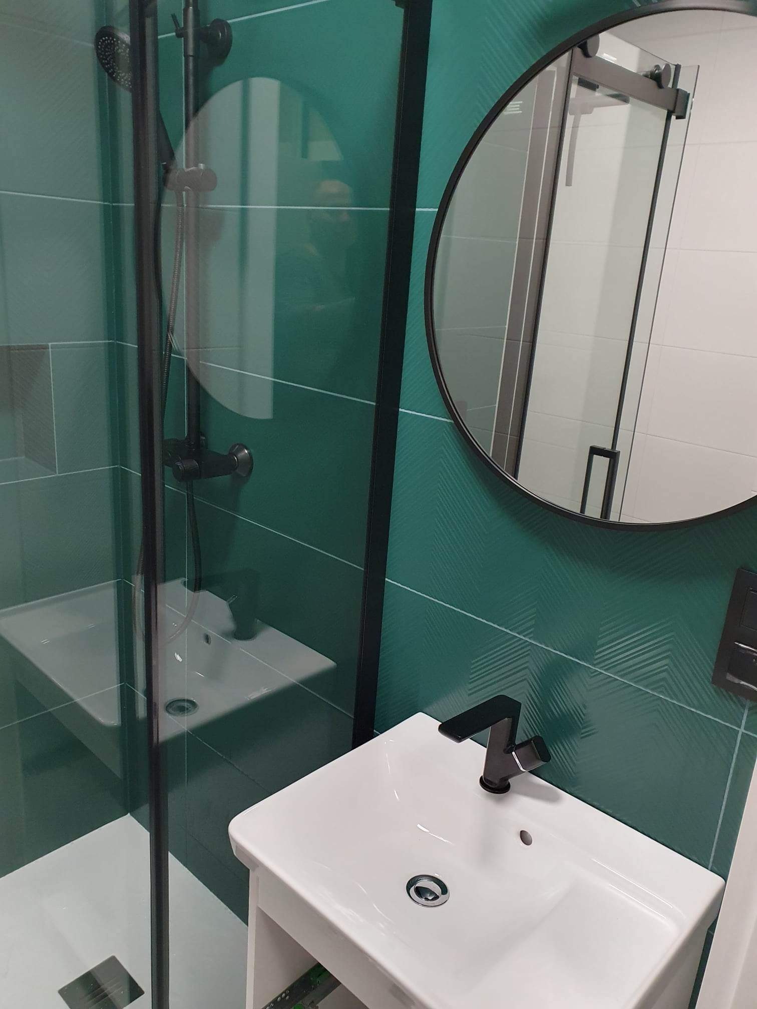 Nowoczesna łazienka z kabiną prysznicową, umywalką nablatową z czarną armaturą i okrągłym lustrem na ścianie z zielonymi płytkami w chevron.