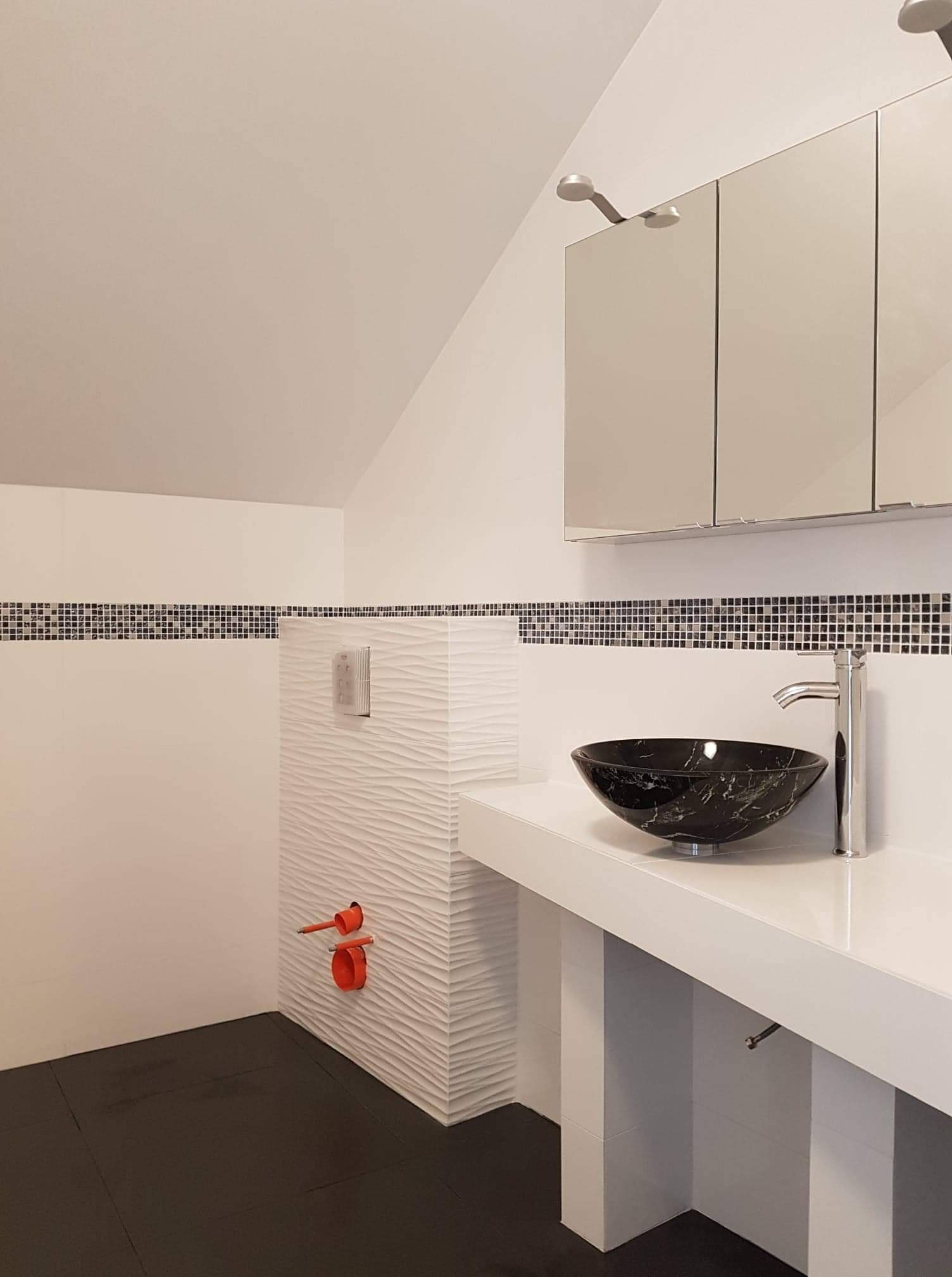 Nowoczesna łazienka na poddaszu z czarną umywalką w kształcie misy, chromowaną baterią, białą szafką z lustrami, mozaiką na ścianie i podwieszanym WC z białą, teksturowaną obudową i czerwonymi rurami.