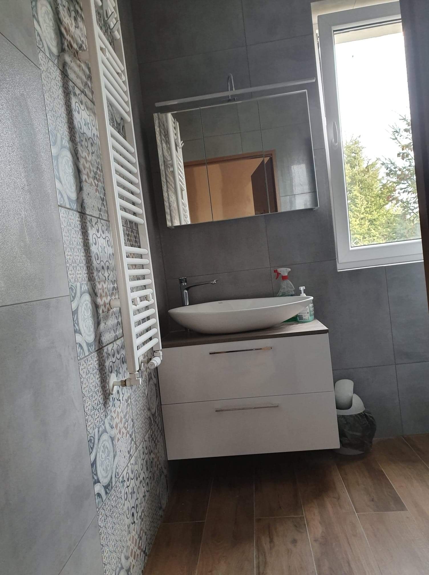 Wykończona łazienka z szarymi płytkami, wzorzystą ścianą, białą umywalką nablatową na szafce z szufladami, grzejnikiem drabinkowym i lustrem w szafce.