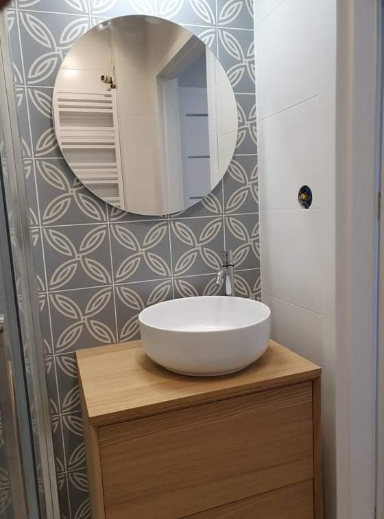 Nowoczesna łazienka z okrągłym lustrem, umywalką nablatową na drewnianej szafce i szarymi płytkami z geometrycznym wzorem.