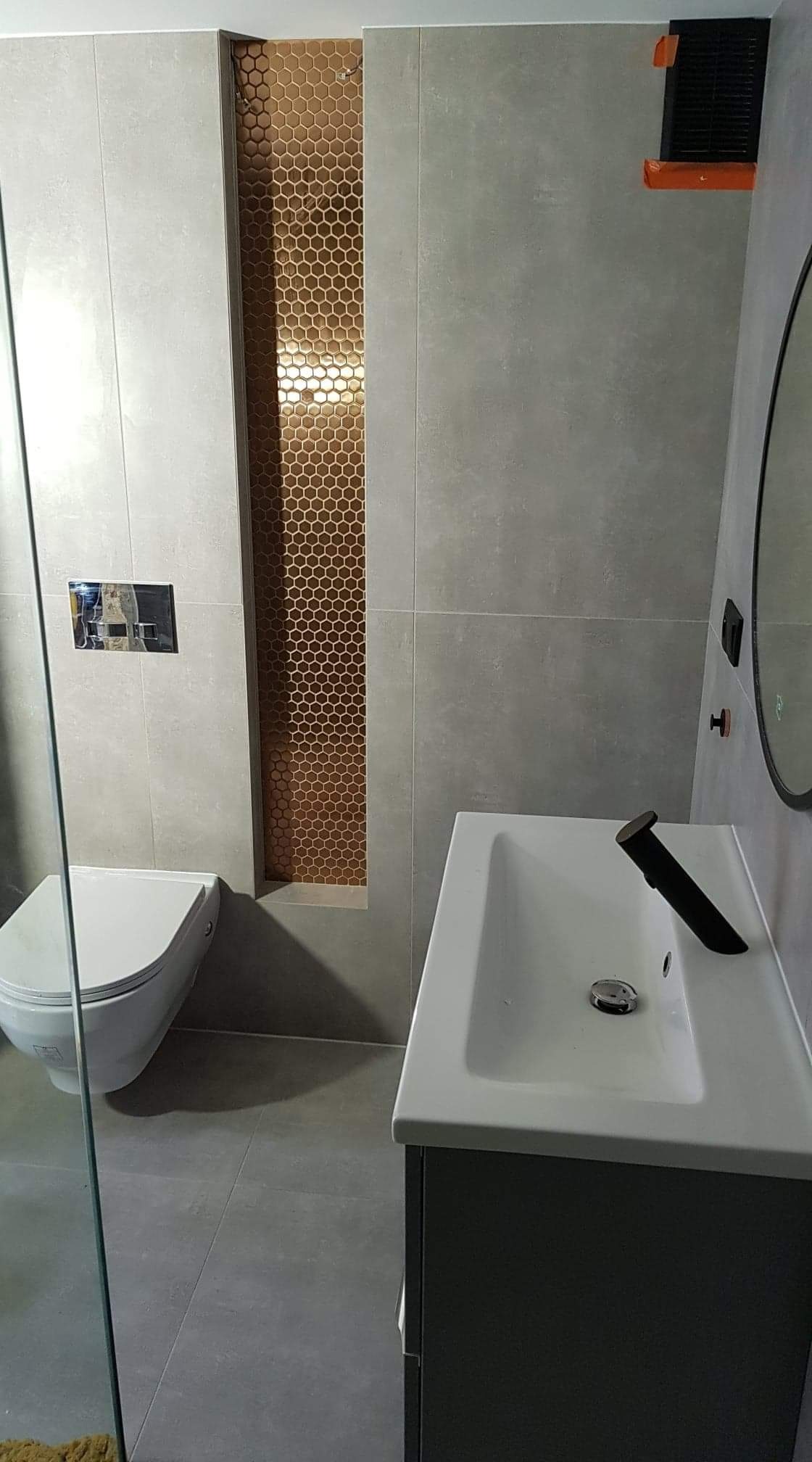 Szara łazienka z toaletą, umywalką z szafką i dekoracyjną wnęką z mozaiką w kształcie plastra miodu.