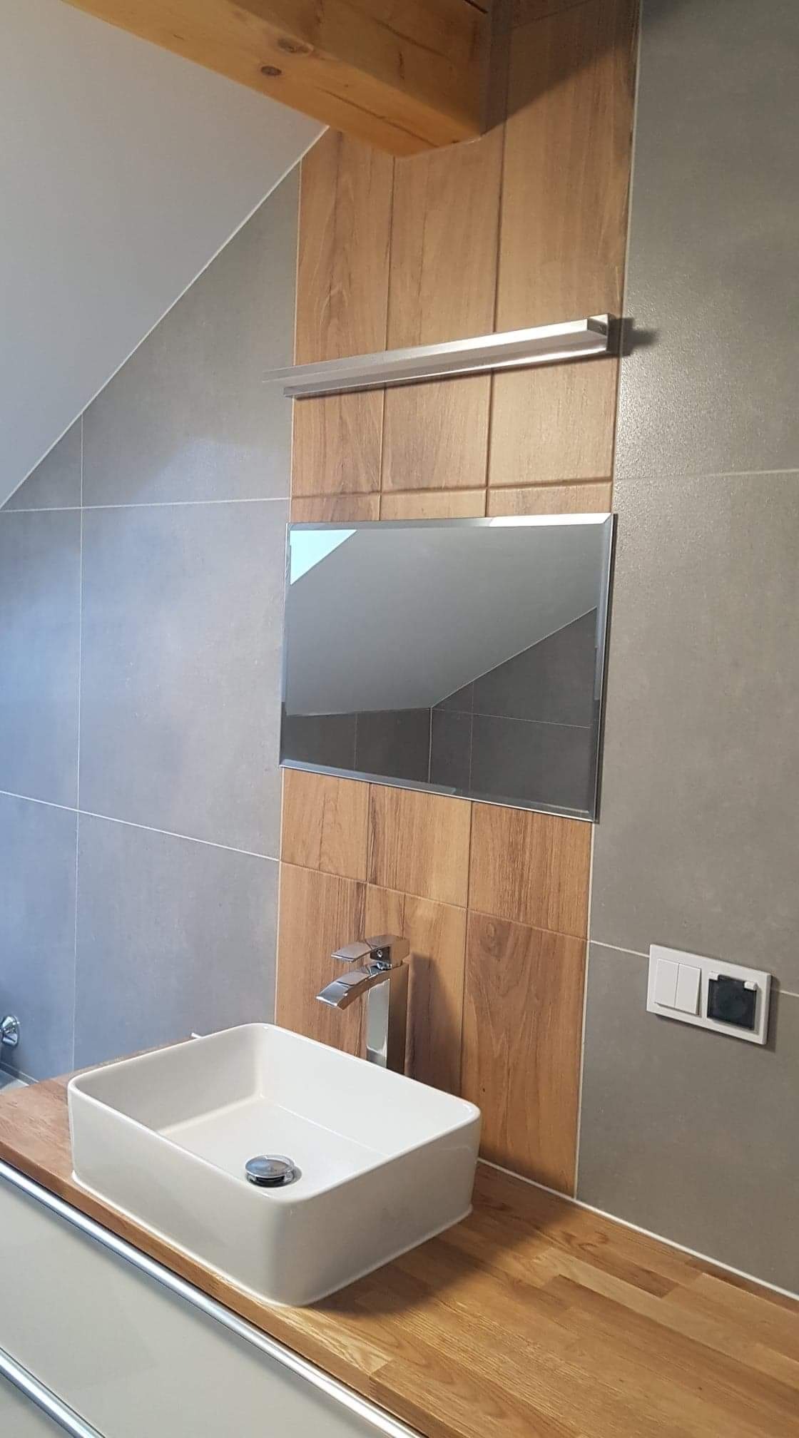 Nowoczesna łazienka z umywalką nablatową, baterią z podwójnym uchwytem, lustrem wpuszczonym w drewnopodobne płytki i szarymi płytkami ściennymi.