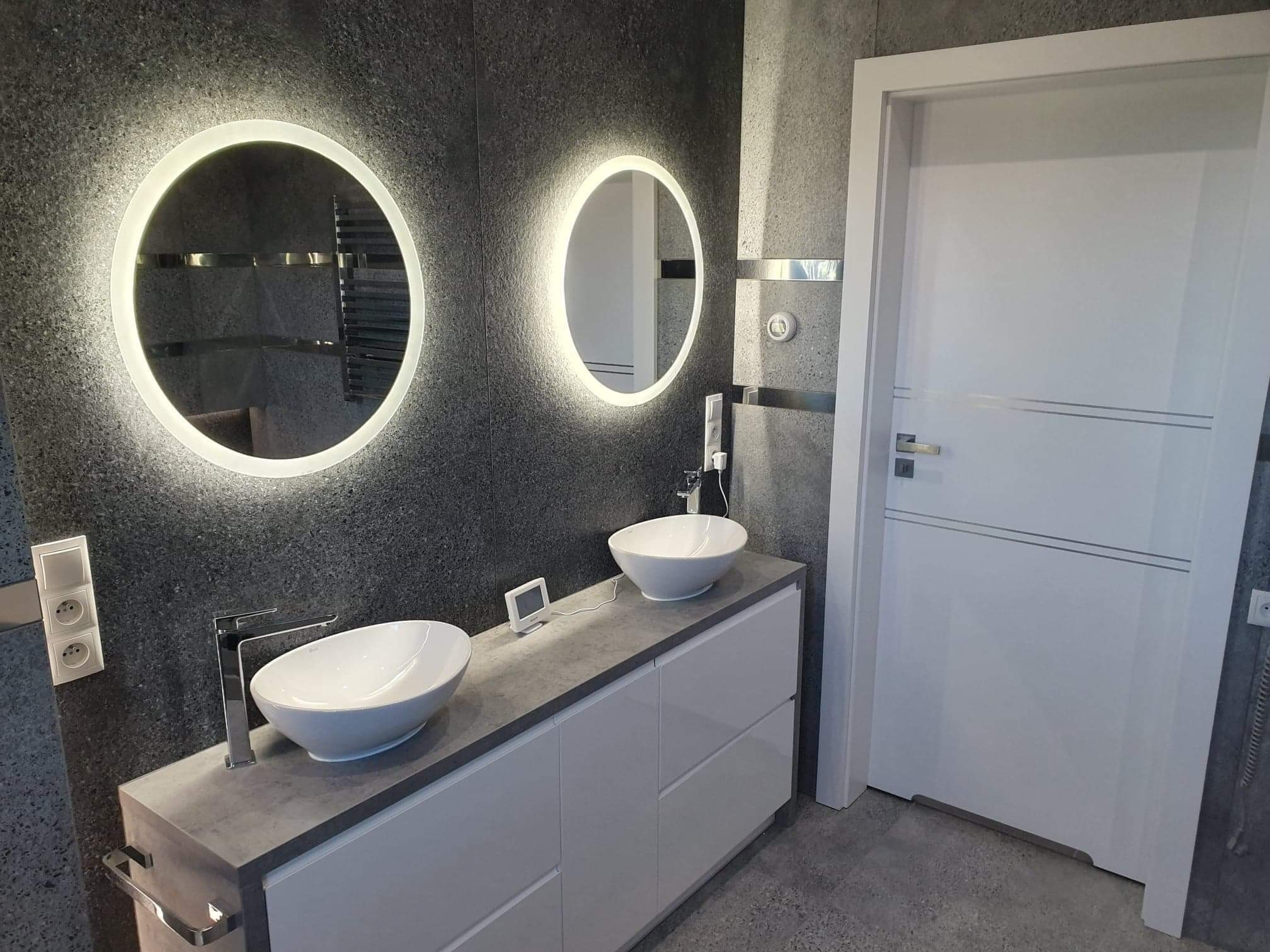 Nowoczesna łazienka z dwoma okrągłymi lustrami z podświetleniem LED, dwiema białymi umywalkami nablatowymi na szarym blacie szafki, szare ściany z fakturą kamienia i białe drzwi z metalowymi...