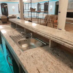 Beżowy blat z kamienia z wbudowanym zlewozmywakiem ze stali nierdzewnej, widoczny w przestronnym wnętrzu w trakcie prac wykończeniowych, z elementami dekoracyjnymi i kolumnami.