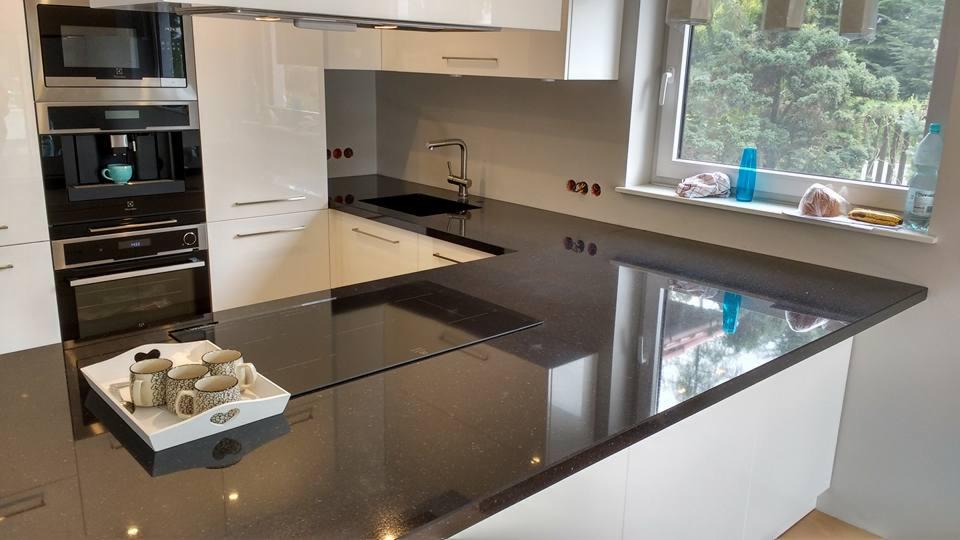 Błyszczący, ciemny blat kamienny w nowoczesnej kuchni z białymi szafkami, w tle okno z widokiem na zieleń i cztery filiżanki na tacy na blacie.