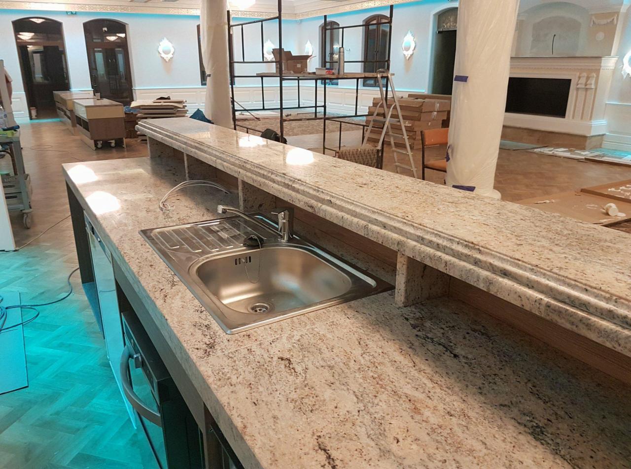Beżowy blat z kamienia z wbudowanym zlewozmywakiem ze stali nierdzewnej, widoczny w przestronnym wnętrzu w trakcie prac wykończeniowych, z elementami dekoracyjnymi i kolumnami.