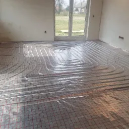 Montaż ogrzewania podłogowego: białe rurki grzewcze ułożone spiralnie na czerwonej siatce w surowym wnętrzu z oknem.