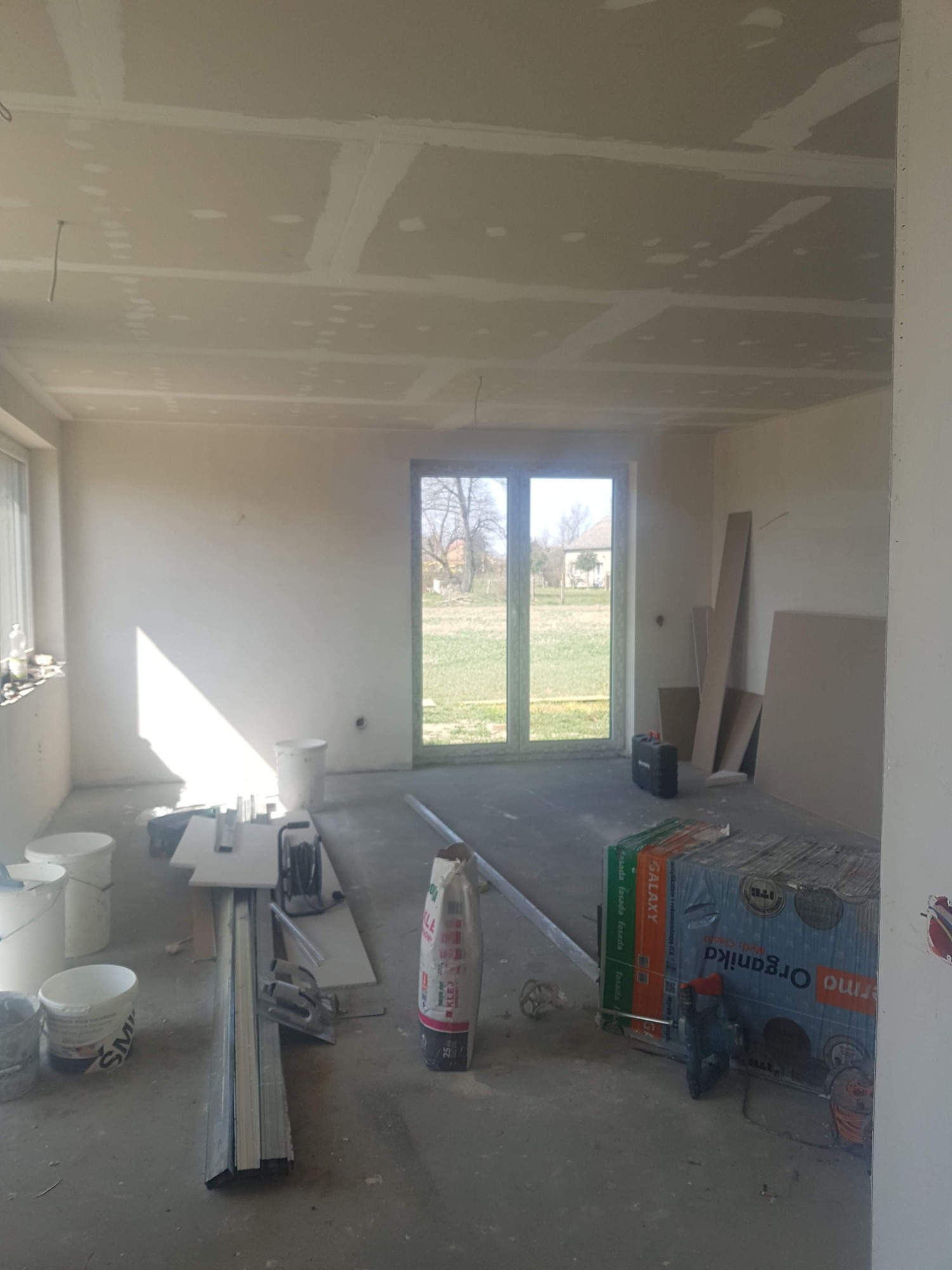 Wnętrze pokoju w trakcie prac wykończeniowych, widoczne płyty na suficie z widocznymi łączeniami, narzędzia i materiały budowlane na podłodze, okno z widokiem na zewnątrz.