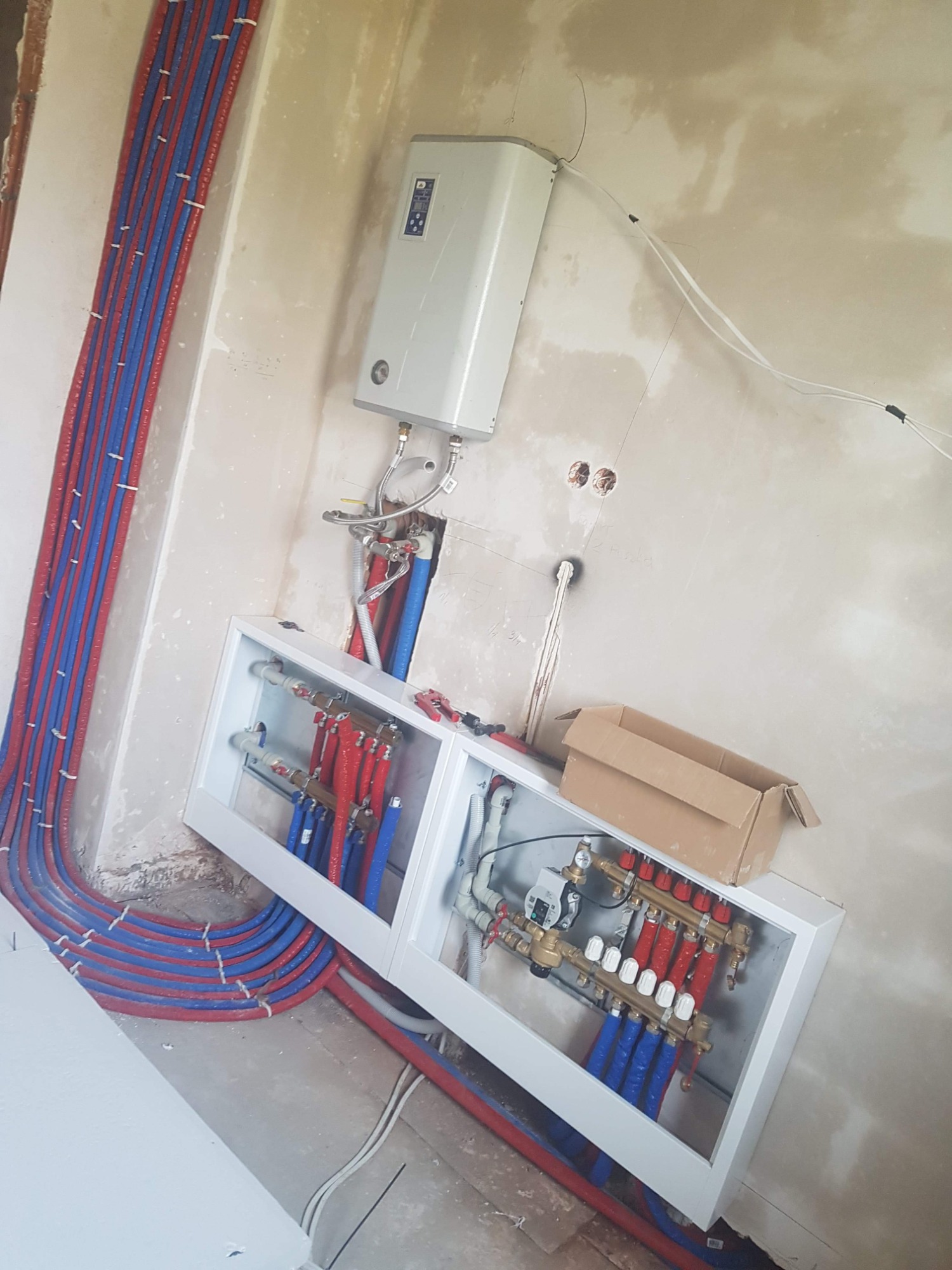 Rozdzielacz instalacji centralnego ogrzewania z licznymi rurami w kolorze czerwonym i niebieskim, zamontowany na ścianie w surowym wnętrzu, z widocznym bojlerem elektrycznym i kartonem obok.
