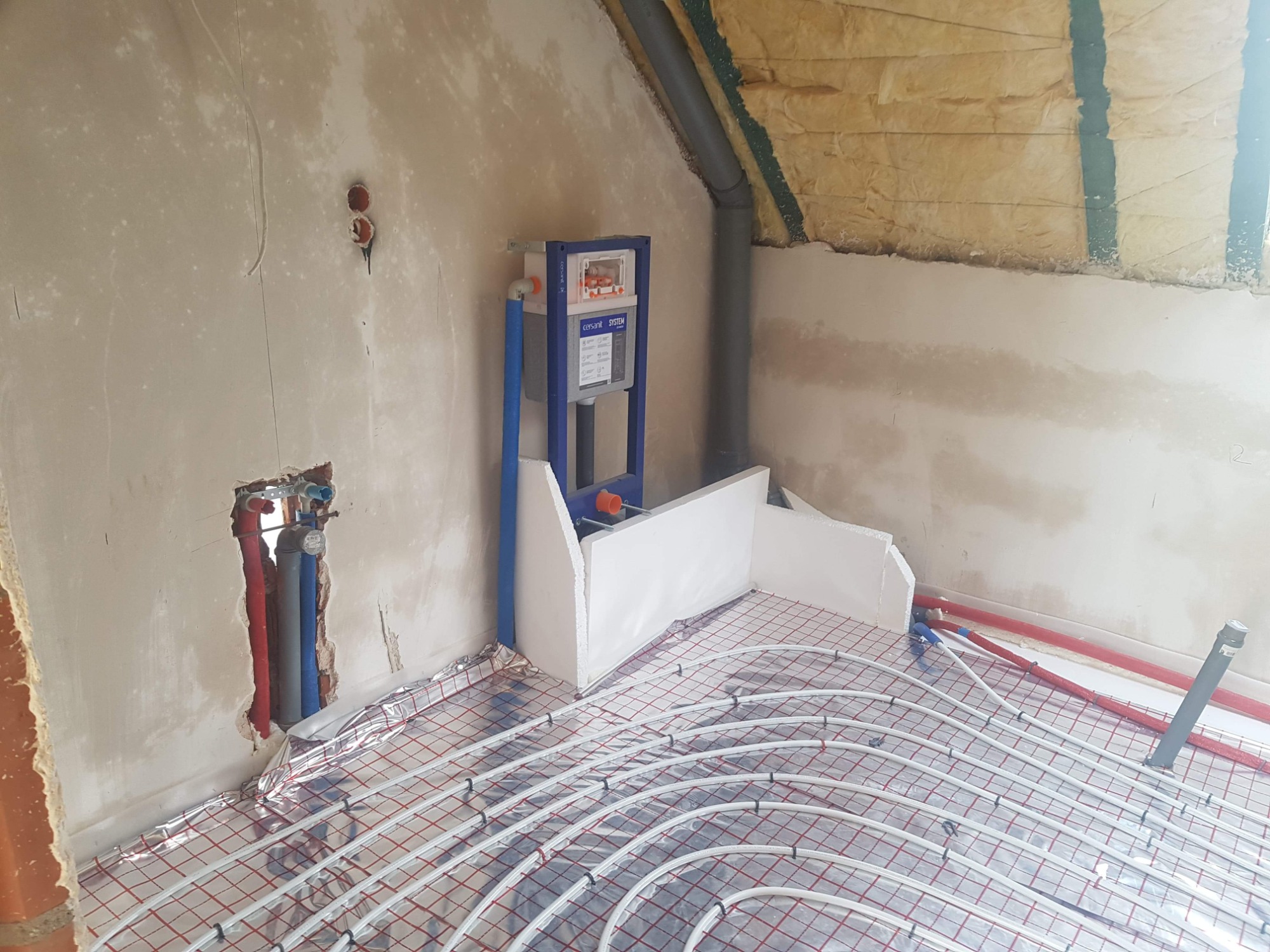 Instalacja ogrzewania podłogowego w pomieszczeniu na poddaszu, widoczne rury grzewcze na folii aluminiowej, stelaż podtynkowy WC oraz ocieplenie skośnego dachu wełną mineralną.