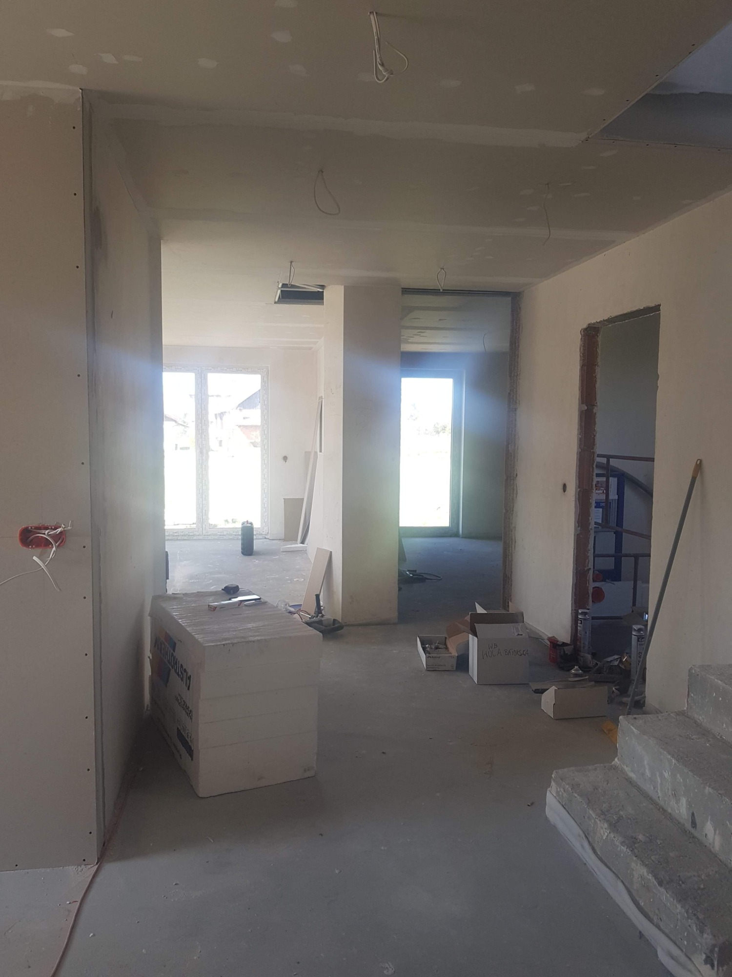 Wnętrze domu w trakcie prac wykończeniowych: widoczne ściany z zamontowanymi płytami, przygotowane schody, okno w głębi oraz materiały budowlane na podłodze.