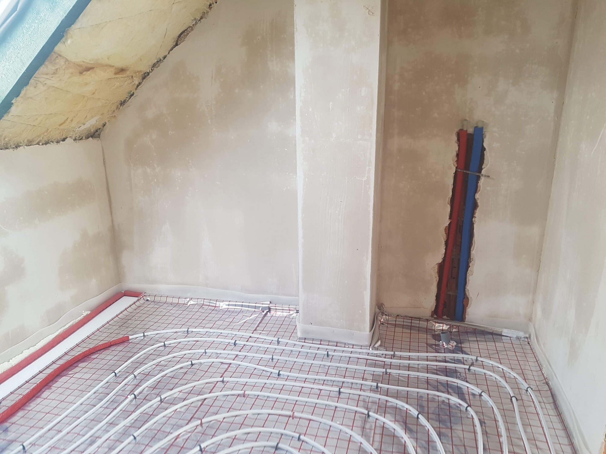 Poddasze w trakcie prac wykończeniowych: izolacja skośnego dachu wełną mineralną, ściany w stanie surowym po szpachlowaniu, instalacja ogrzewania podłogowego z rur na folii z siatką, widoczne rury...