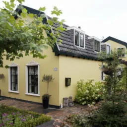 Żółty dom jednorodzinny z ciemną dachówką, białymi ramami okiennymi z dekoracyjnymi elementami i zadbanym ogrodem z kwitnącymi hortensjami. Widoczny numer domu '47' i skrzynka na listy.