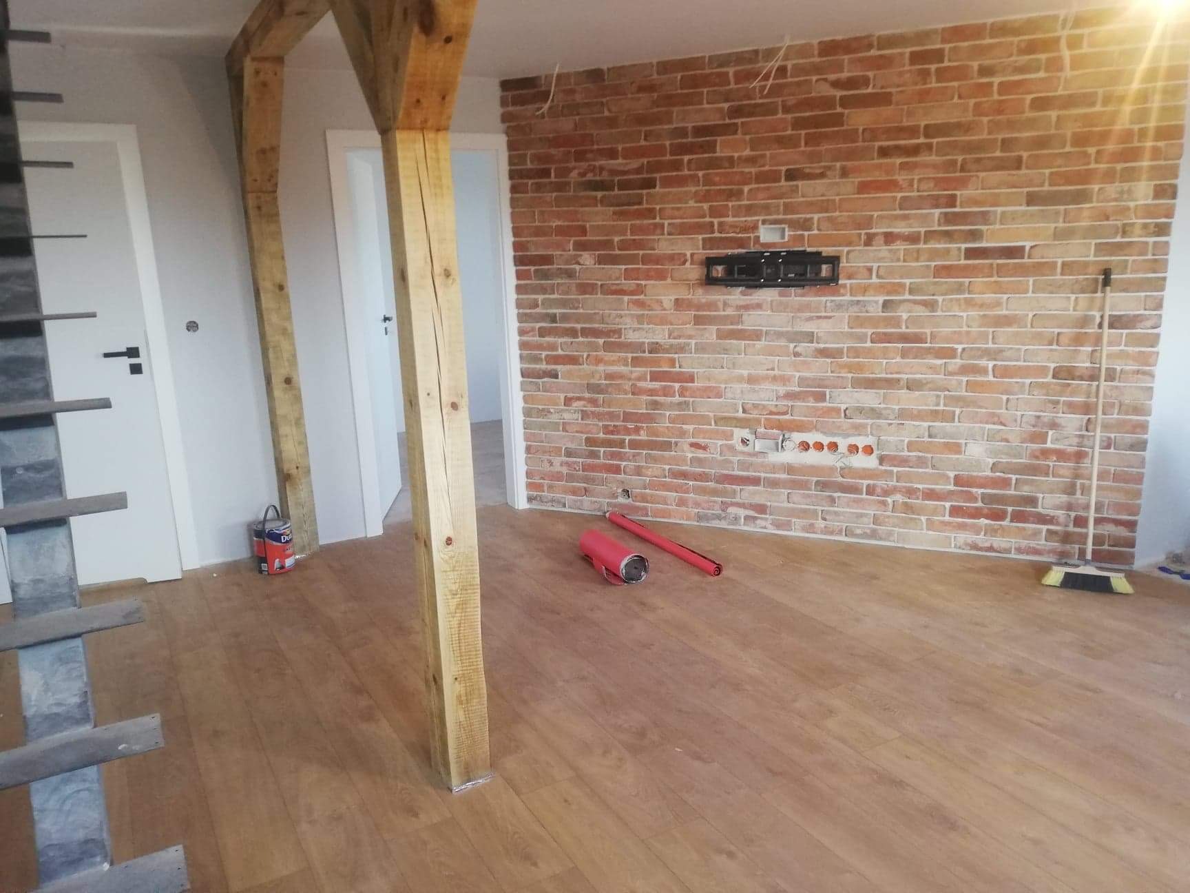 Pokój po remoncie z drewnianą podłogą, ceglaną ścianą, drewnianymi belkami, widoczne narzędzia malarskie i puszka z farbą.