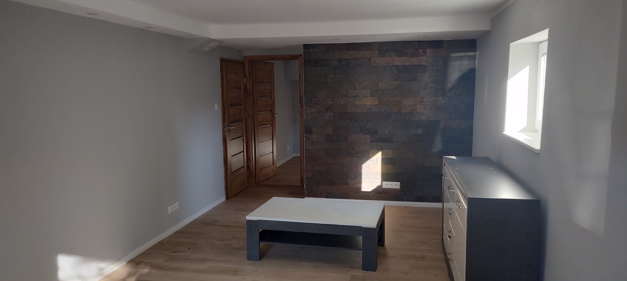 Szare ściany, drewniana podłoga i okładzina z kamienia dekoracyjnego na ścianie. Widoczne drewniane drzwi i komoda. Jasne wnętrze po remoncie.