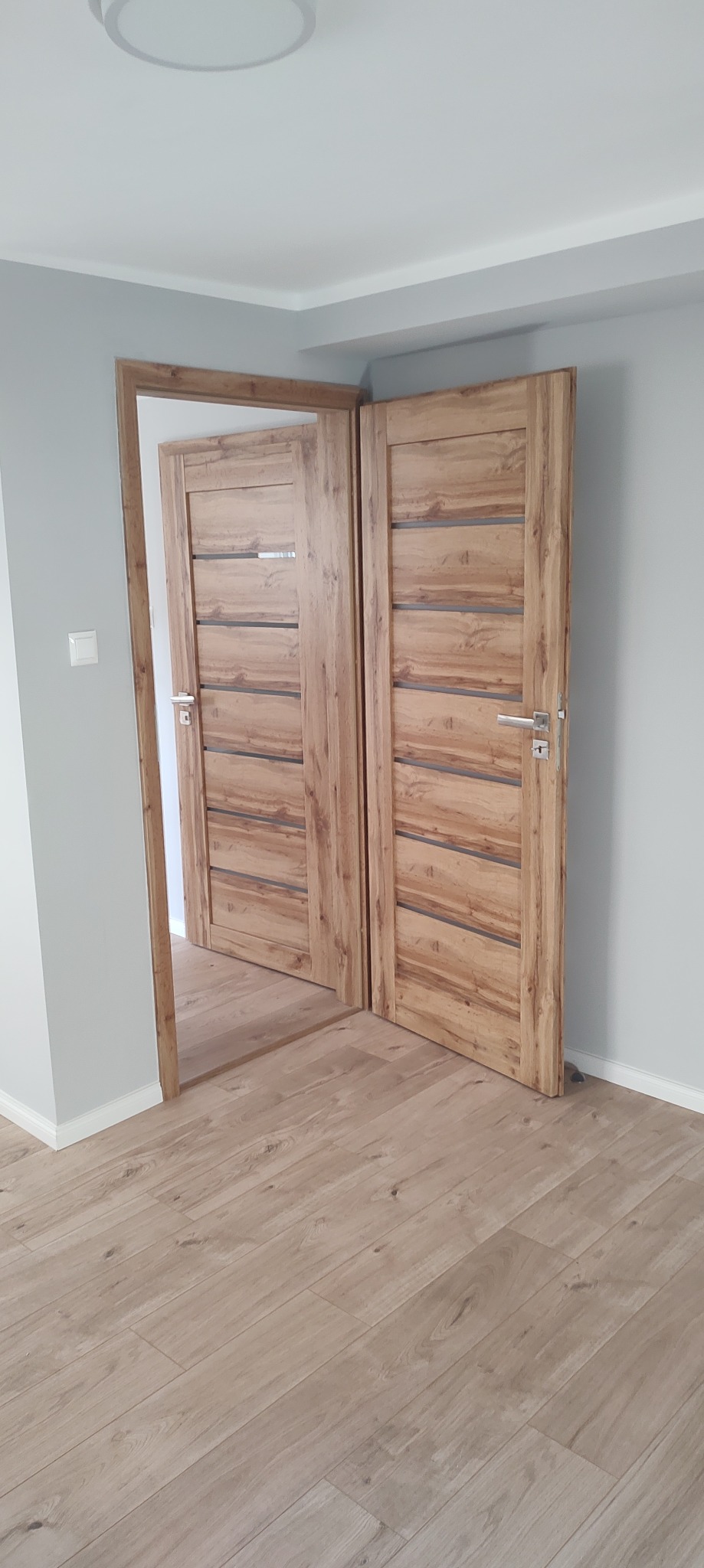 Dębowe drzwi wewnętrzne z przeszkleniami, zamontowane w nowym domu w Bartągu. Widoczna podłoga z paneli i szare ściany. Drzwi otwarte, ukazują wnętrze.