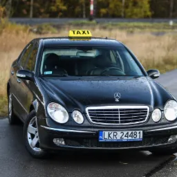 Czarny Mercedes taxi z żółtym szyldem 'TAXI' na dachu, zaparkowany na mokrej drodze w jesiennej scenerii.