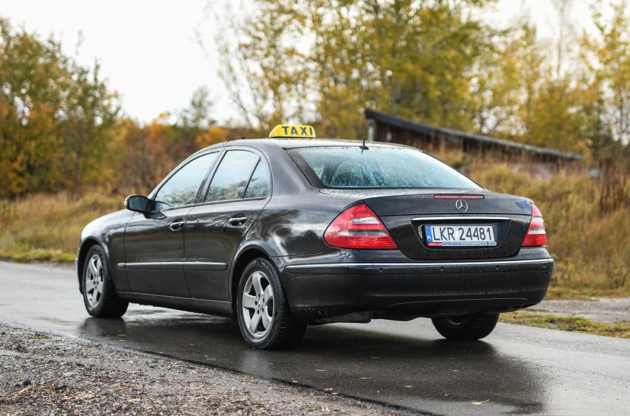 Czarna taksówka Mercedes z żółtym oznaczeniem na dachu, widoczna z tyłu na mokrej drodze w jesiennym krajobrazie.