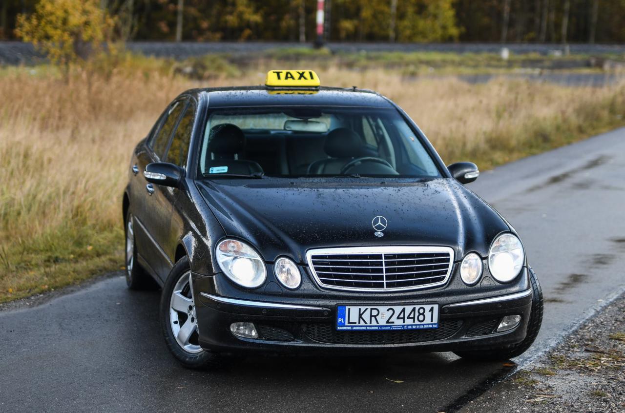 Czarny Mercedes taxi z żółtym szyldem 'TAXI' na dachu, zaparkowany na mokrej drodze w jesiennej scenerii.
