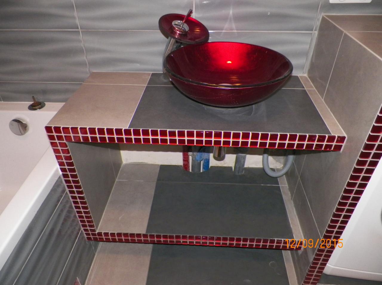 Szklana, czerwona umywalka w kształcie misy na blacie z szarych i beżowych płytek, wykończonymi bordową mozaiką, widoczne rury odpływowe, szare płytki na ścianie w tle oraz fragment wanny po lewej...