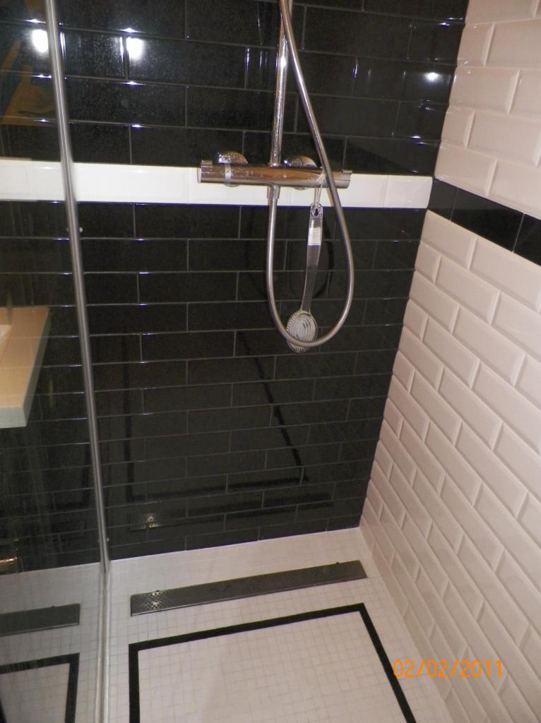 Nowoczesna kabina prysznicowa z czarnymi błyszczącymi płytkami na ścianie i białymi płytkami typu metro na sąsiedniej ścianie, chromowana armatura i odpływ liniowy.