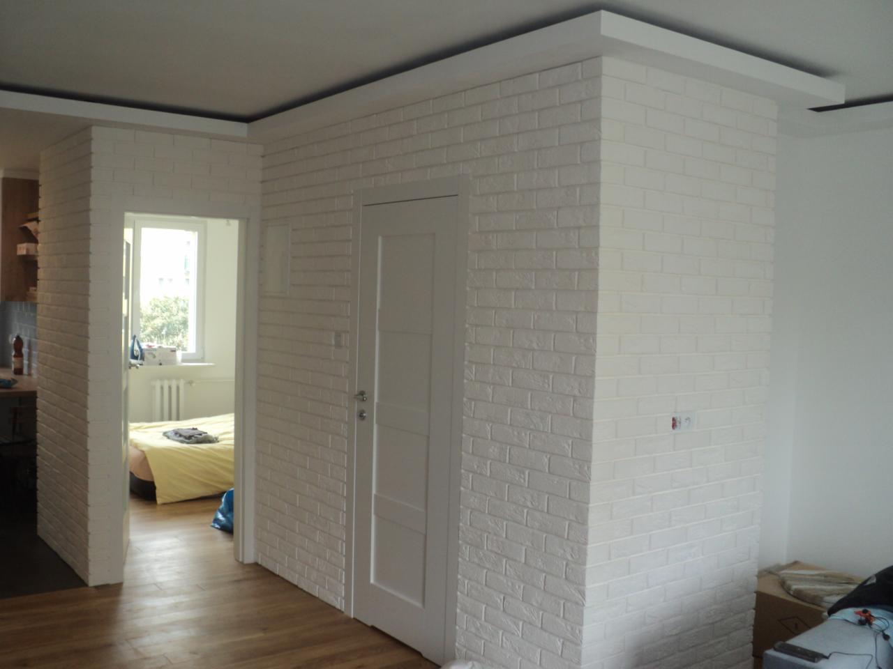 Ściana wykończona białą cegłą dekoracyjną, z widocznymi drzwiami i fragmentem pokoju z żółtą pościelą w tle.