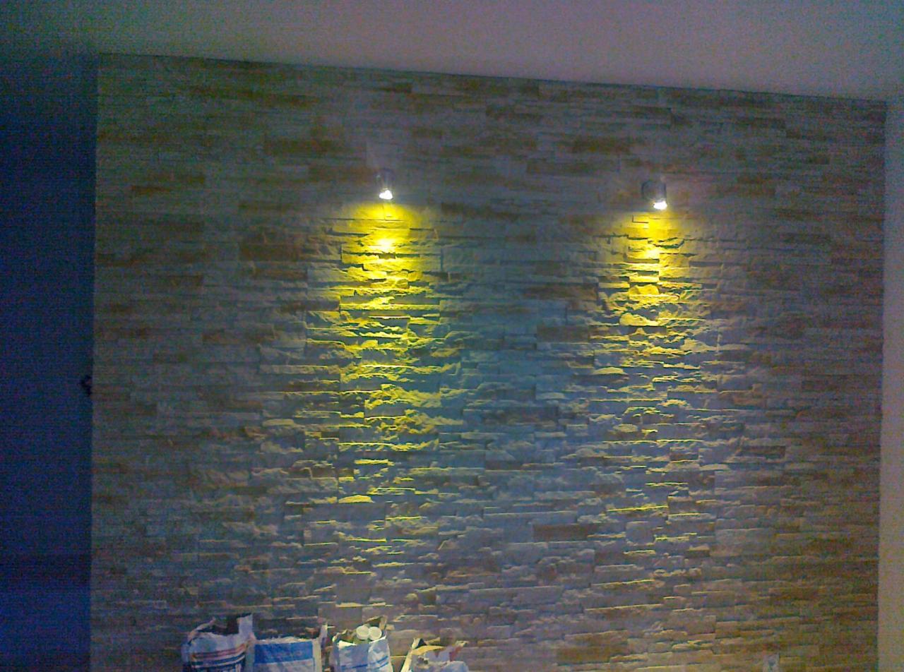 Ściana wykończona kamieniem dekoracyjnym, oświetlona dwoma punktowymi lampami, z workami materiałów budowlanych u dołu.
