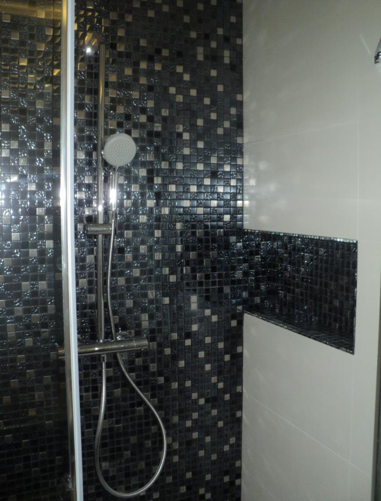 Wykończona kabina prysznicowa z błyszczącą, czarno-srebrną mozaiką kwadratową i wnęką wyłożoną tym samym materiałem, kontrastująca z gładką, białą ścianą.