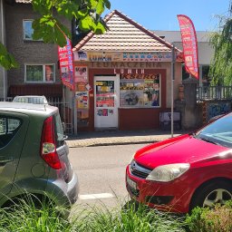 LUMEN - Fasada punktu usługowego 'Lumen Ksero' z ofertą tłumaczeń, ksero i pieczątek, widoczna zza zieleni i zaparkowanych aut, w słoneczny dzień.