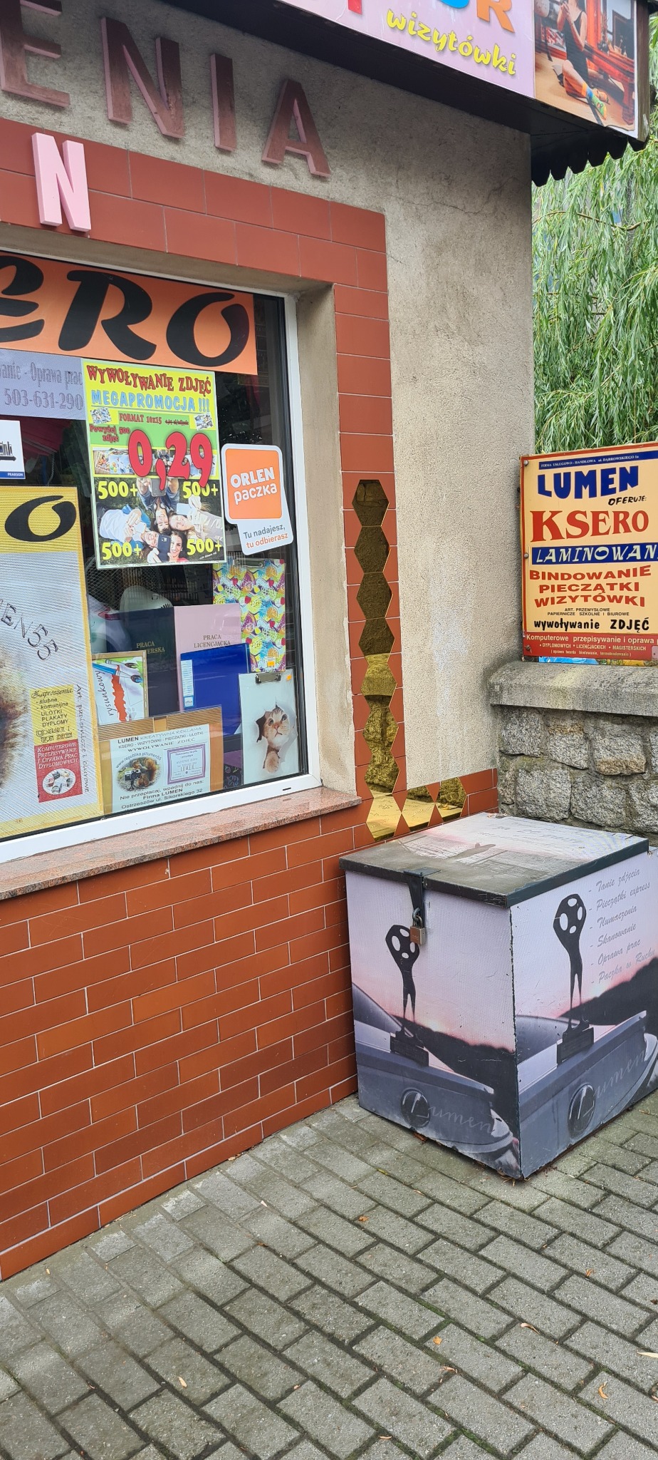 Fasada punktu usługowego Lumen z reklamami wywoływania zdjęć, ksero i wizytówek. Na chodniku stoi oklejony pojemnik z logo firmy i hasłami reklamowymi.