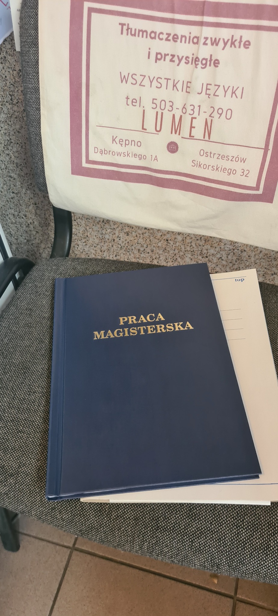 Oprawiona praca magisterska z napisem w kolorze złotym leży na krześle obok torby z logo biura tłumaczeń Lumen, oferującego tłumaczenia zwykłe i przysięgłe.