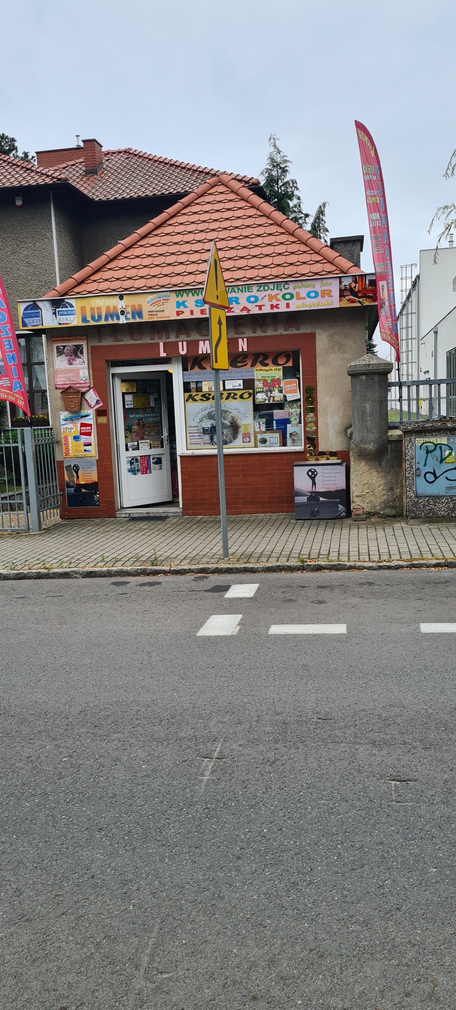 Fasada punktu usługowego Lumen Ksero z ofertą wywoływania zdjęć, ksero i pieczątek, widoczna z ulicy, z reklamami na oknach i chodniku.