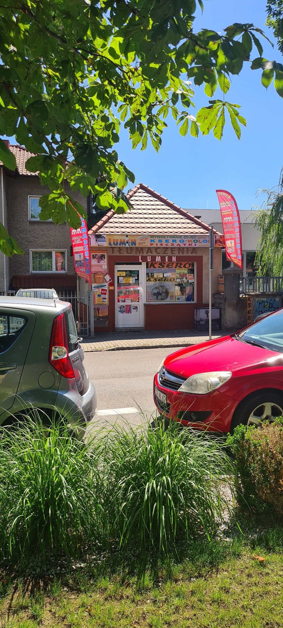 Fasada punktu usługowego 'Lumen Ksero' z ofertą tłumaczeń, ksero i pieczątek, widoczna zza zieleni i zaparkowanych aut, w słoneczny dzień.