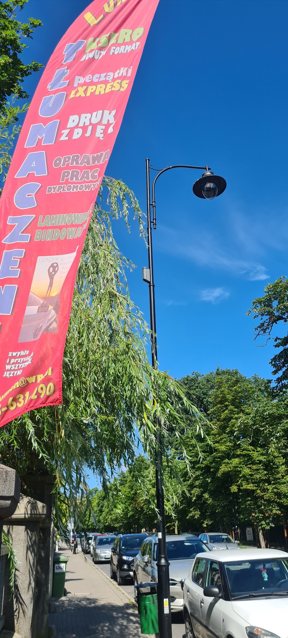 Czerwony, pionowy baner reklamowy z napisem 'Tłumaczenia' i ofertą usług poligraficznych na tle błękitnego nieba i zieleni drzew. Widoczny fragment ulicy z samochodami.