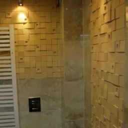 Nowoczesna łazienka z beżowymi płytkami 3D na ścianie, podświetlona punktowym oświetleniem, z widocznym grzejnikiem drabinkowym i podwieszaną toaletą.