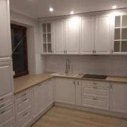 Jasna kuchnia z białymi frontami szafek w stylu klasycznym, drewniany blat, płytki na ścianie, okno z ciemną ramą i drewniana podłoga.