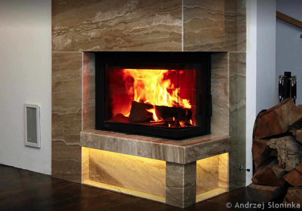 Nowoczesny kominek z obudową z kamienia naturalnego o falistej strukturze, podświetlony od dołu ciepłym światłem, z paleniskiem wypełnionym płomieniami i drewnem, obok ułożone drewno opałowe.