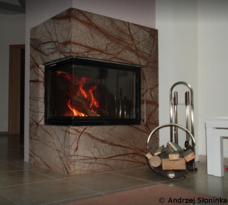 Narożny wkład kominkowy z obudową z kamienia o wyraźnym, żyłkowatym wzorze, w tle widoczny kosz na drewno i akcesoria kominkowe.