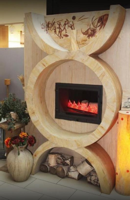 Nowoczesny kominek z obudową z jasnego drewna o unikalnym kształcie z motywem prehistorycznym, imitacja płomieni wewnątrz, ułożone drewno pod spodem, wazon z kwiatami obok.