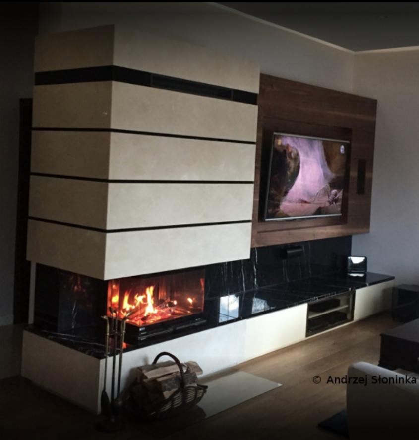 Nowoczesny, narożny piec z paleniskiem i obudową z jasnego kamienia z czarnymi pasami, zlokalizowany w Starym Górniku, z telewizorem w drewnianej obudowie nad nim i drewnianą podłogą.