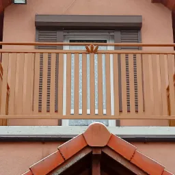 Balkon z drewnianą balustradą w kolorze brązowym, widok od dołu, z fragmentem dachu i oknem w tle.