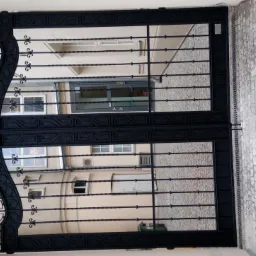 Czarna, metalowa brama z ozdobnymi elementami w kształcie wolut, umieszczona w łukowatym przejściu, widok na dziedziniec z budynkami o jasnej elewacji i brukowaną nawierzchnią.