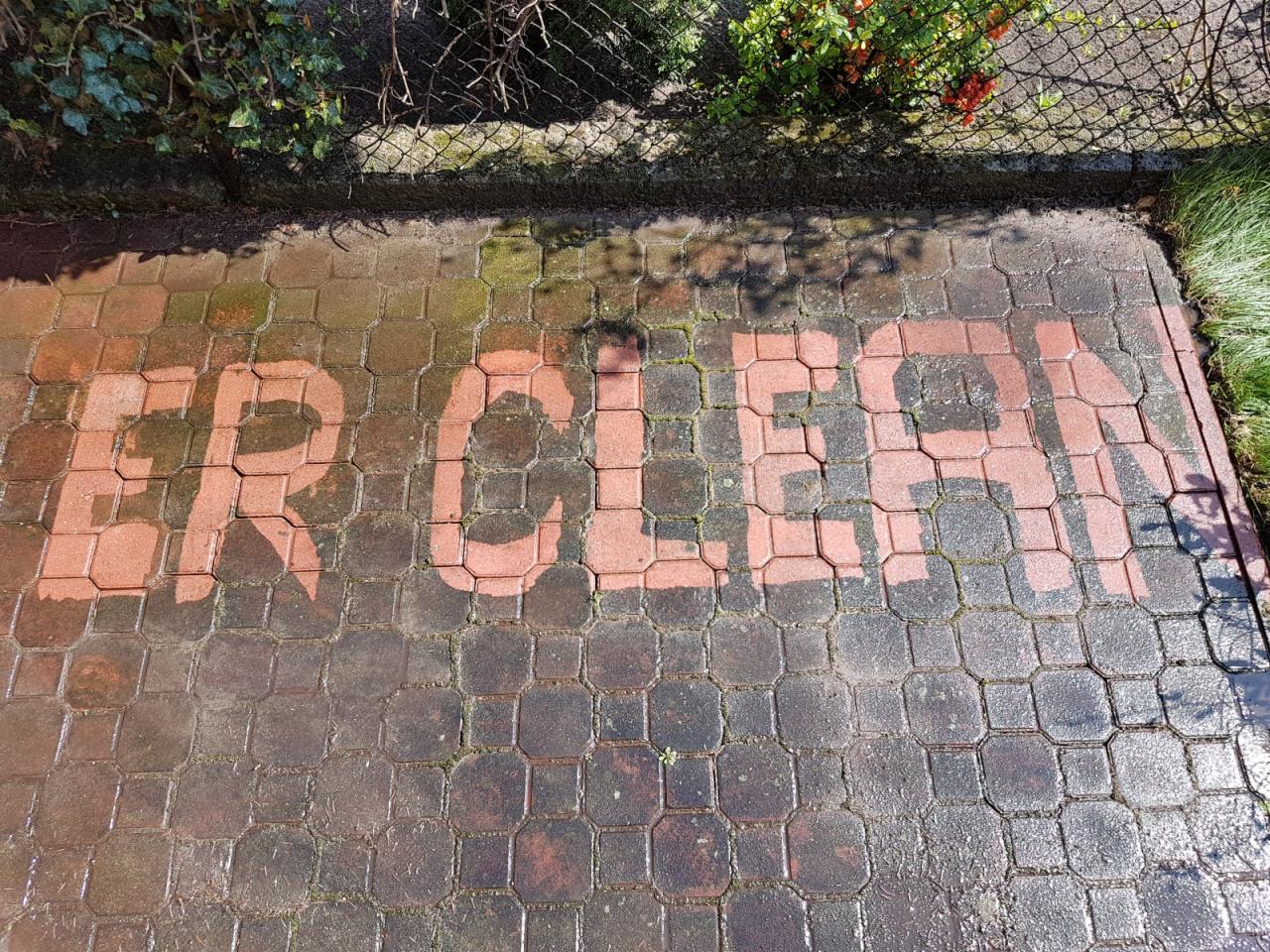 Częściowo umyta kostka brukowa z napisem 'ER CLEAN', widoczne różnice w kolorze i wilgotności, zarośla w tle.