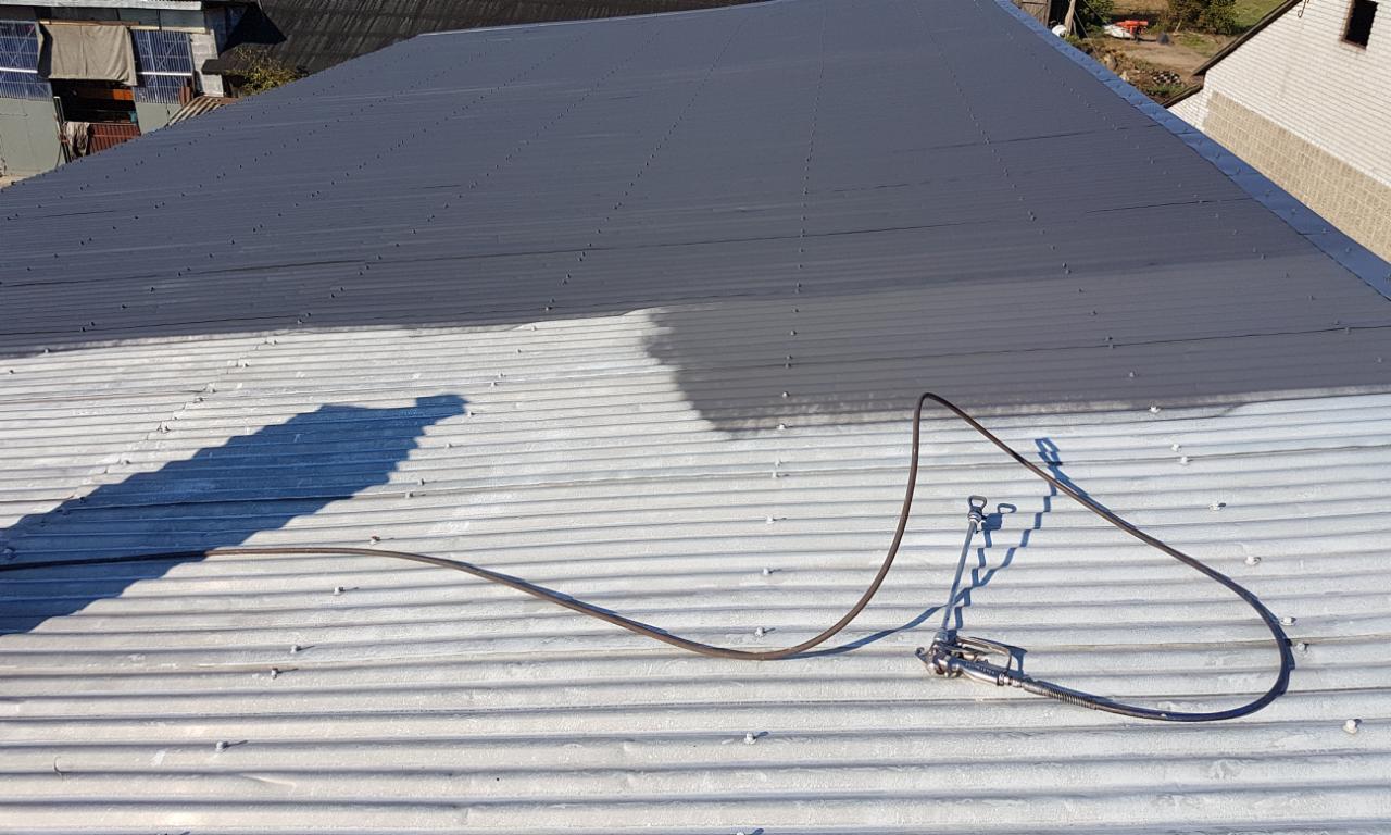 Częściowo pomalowany na szaro blaszany dach z widocznym pistoletem natryskowym i wężem, zarys budynku w tle.