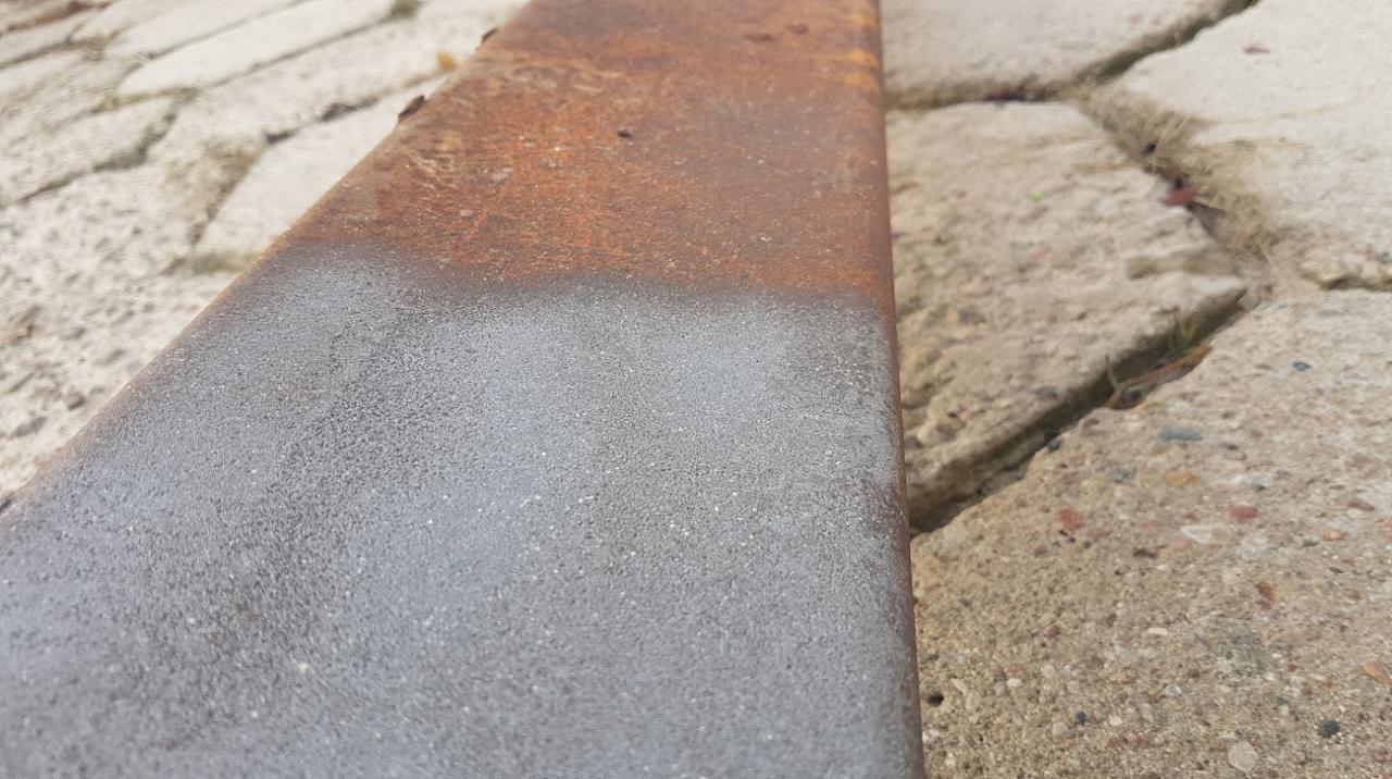 Metalowy element, częściowo oczyszczony z rdzy, leżący na betonowej kostce brukowej. Widoczne przejście od zardzewiałej powierzchni do oczyszczonej, szarej metalicznej barwy.