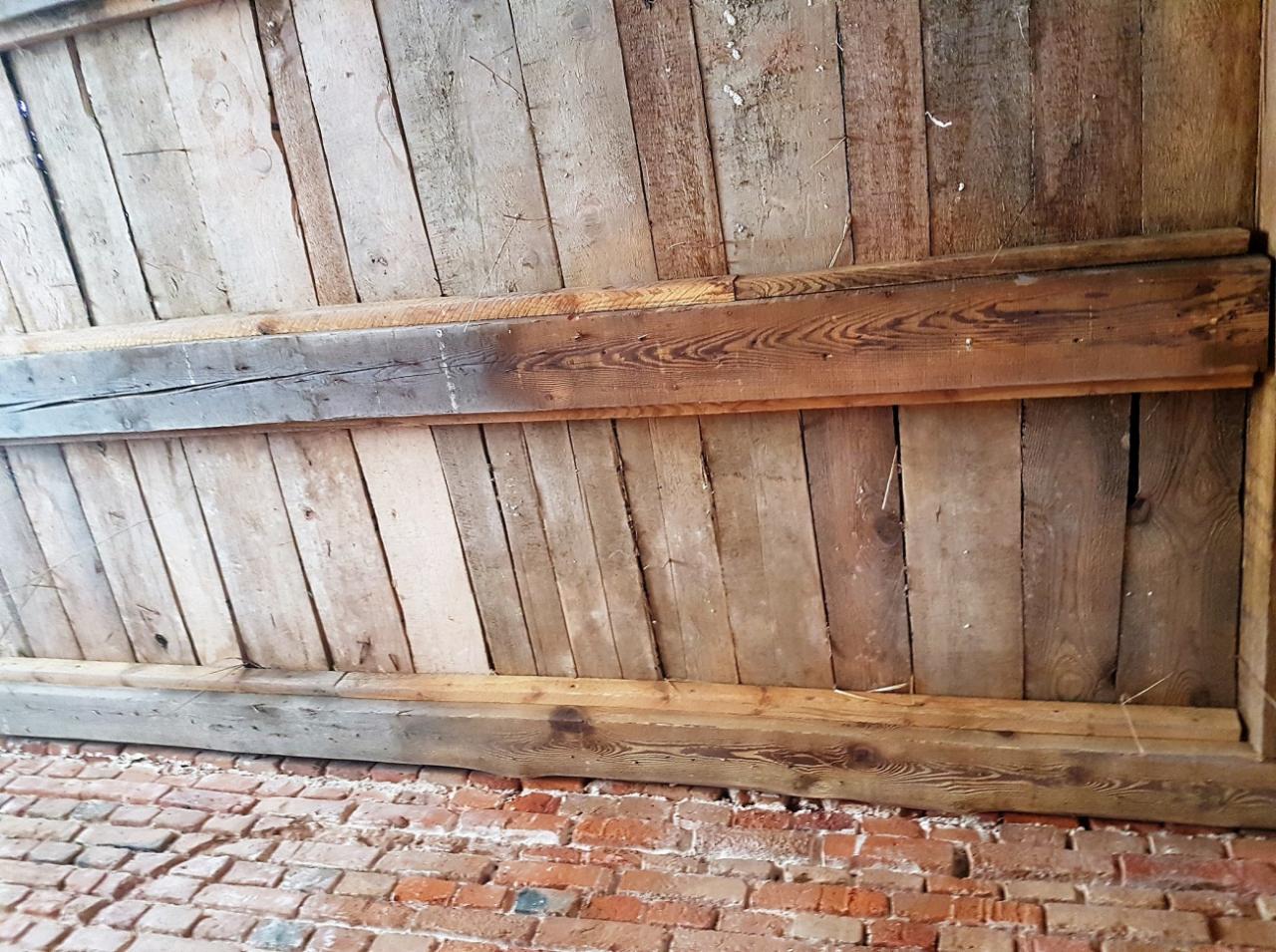 Drewniany strop z belkami i ceglana ściana, widoczne ślady użytkowania i zabrudzenia, ukośny kąt ujęcia.
