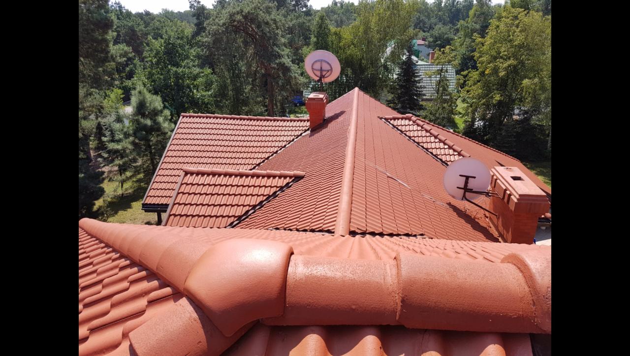 Czerwony dach z dachówki ceramicznej po renowacji, widoczne kominy i anteny satelitarne, tło stanowią drzewa.